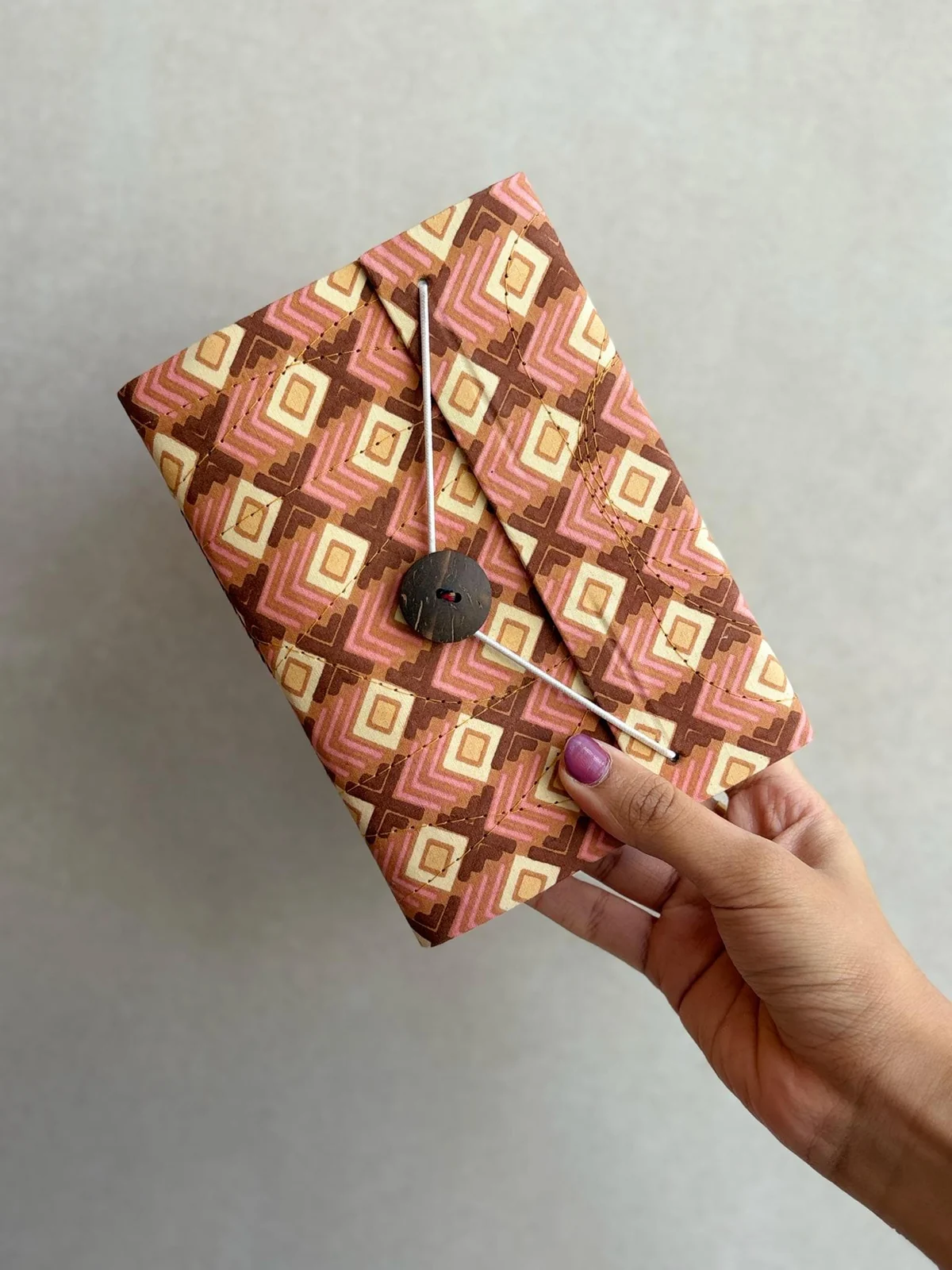 A hand block printed
        notebook in amber, rust and cream diamonds, closed with a coconut button and
        held in one hand.